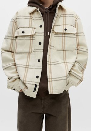 Hombre con chaqueta abotonada a cuadros en crema y beige sobre sudadera marrón con capucha y manos en los bolsillos, combinado con pantalones marrón oscuro.