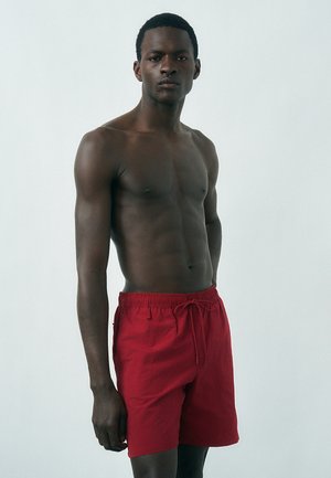 Άνδρας με σκουρόχρωμη επιδερμίδα χωρίς πουκάμισο, φορεμένος με κόκκινα σορτς με κορδόνι, στέκεται μπροστά σε έναν απλό ανοιχτό γκρι φόντο και κοιτάζει μπροστά.