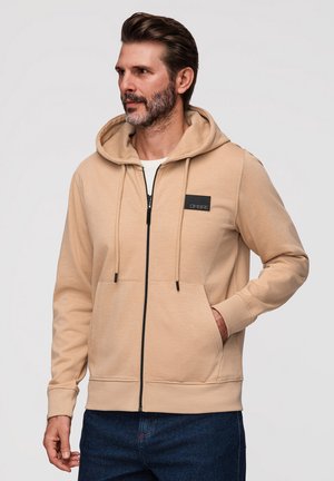 Homme barbu portant un sweat à capuche zippé beige avec poches avant et un jean bleu foncé, debout devant un fond clair uni.