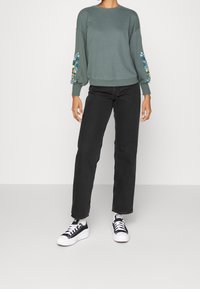 Sweatshirt gris avec broderie florale sur les manches, associé à un jean droit noir et des baskets en toile noir et blanc.