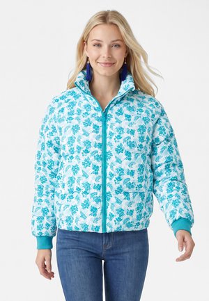Lächelnde Frau mit langen blonden Haaren, die eine blaue, geblümte Steppjacke, blaue Jeans und dunkelblaue Quastenohrringe trägt, steht vor weißem Hintergrund.