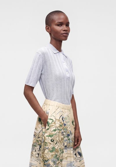 Femme aux cheveux courts portant une chemise à manches courtes gris clair avec col et une jupe beige avec un motif de paysage vert et bleu.