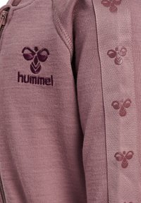 Lilla sweatshirt med lynlås, der har et broderet logo på brystet og mønstret sidebånd. Blød tekstur og afslappet stil.