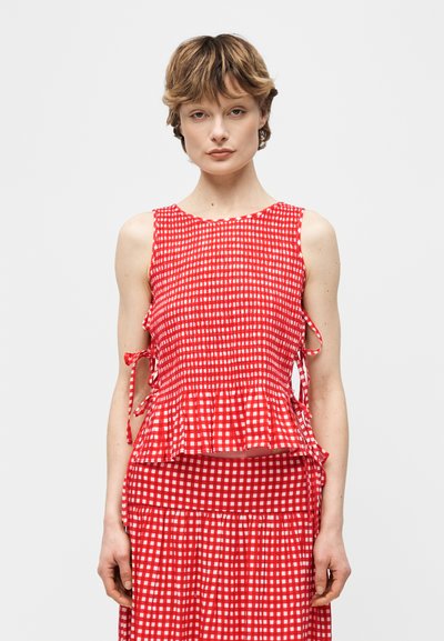 Femme aux cheveux courts portant un haut sans manches à carreaux rouges et blancs avec des liens sur les côtés et une jupe assortie, debout devant un fond clair uni.
