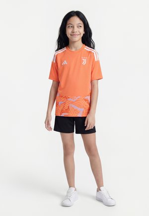 Ung pige med langt sort hår iført en orange Juventus fodboldtrøje, sorte shorts og hvide sneakers, står og smiler.