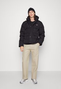 Giacca nera imbottita con cappuccio, chiusura con zip e bottoni a pressione, tasche frontali, abbinata a pantaloni beige e sneakers, su uno sfondo neutro.