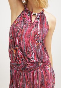 Vrouw in een mouwloze jurk met een rood en roze gedraaid patroon, een geraffineerde taille en een halternek met een rechthoekige metalen sluiting.