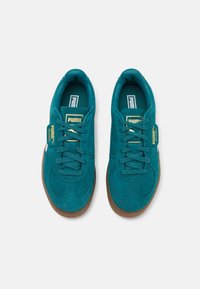 Teal semskinn sneakers med snøring, brun gummisåle og gull Puma-logo på tungen og siden. Minimalistiske geometriske broderidetaljer.