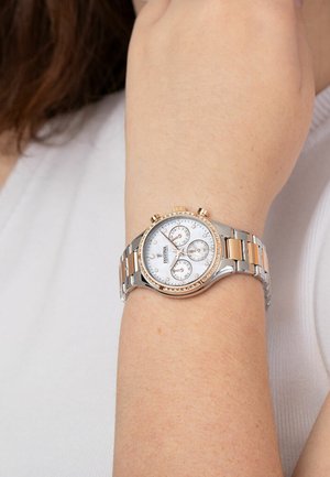 Montre en métal argenté et doré avec un cadran rond blanc, trois sous-cadrans et des accents en cristal sur la lunette, portée au poignet.