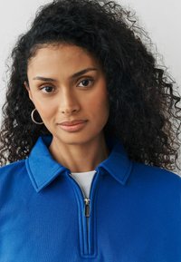 Vrouw met krullend zwart haar, draagt zilveren oorbellen en een blauwe ritssluiting bovenkleding voor een effen lichte achtergrond.