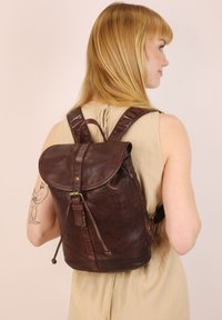 Brązowy skórzany plecak z zaokrągloną klapą, regulowanymi paskami, mosiężnymi elementami oraz teksturowanym wykończeniem. Idealny na codzienne użycie.