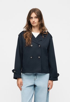 Junge Frau mit langem welligem Haar trägt eine marineblaue zweireihige Jacke und hellblaue Jeans vor einem schlichten weißen Hintergrund.