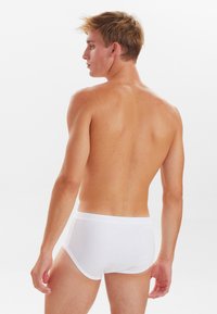 Hvide bomuldsunderbukser med en blød elastik i taljen, tætsiddende design og en glat tekstur. Modellen vises fra siden, bar hud synlig på overkroppen.