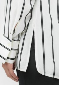 Hvid skjorte med sorte lodrette striber, lange ærmer og en sideslids. Stoffet ser letvægtigt ud med en glat tekstur og knappede manchetter.