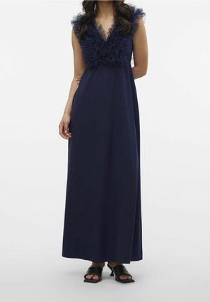 Frau trägt ein ärmelloses, bodenlanges marineblaues Kleid mit gerafftem V-Ausschnitt und schwarze offene Sandaletten mit Absatz, steht vor einfarbigem Hintergrund.