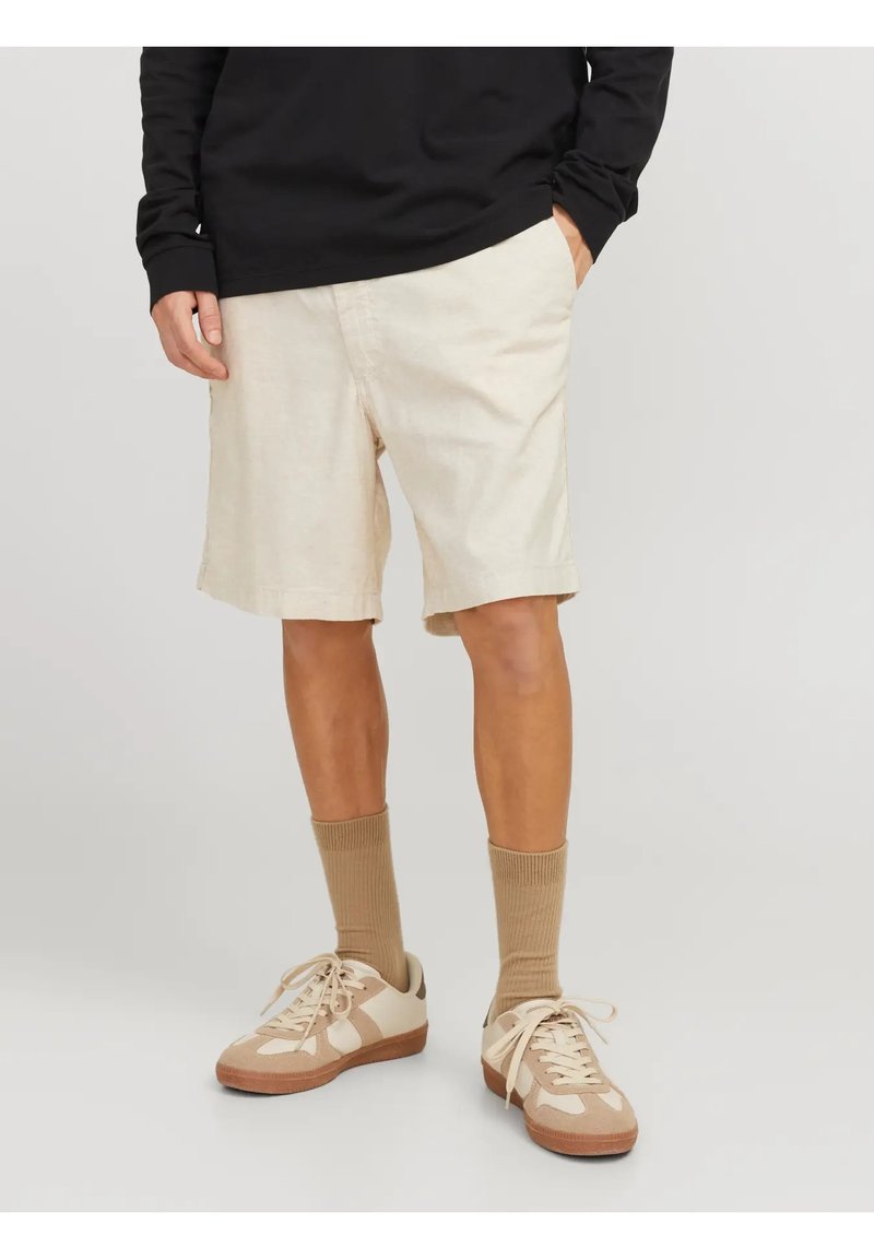 Pantaloni corti in lino beige con tasche laterali, abbinati a sneaker marrone chiaro e calze abbinate. Indossati con una maglietta nera a maniche lunghe.