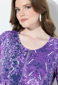 Haut à motif violet avec un col rond et une demi-fermeture éclair en métal argenté. Le tissu présente un design marbré et texturé.