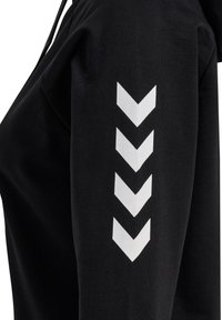 Svart huvtröja med en mjuk textur, med ett vitt chevronmönster tryckt på överarmen. Justerbar huva och avslappnad passform.