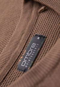 Suéter marrón de punto con una superficie texturizada, que cuenta con un cuello y una etiqueta que dice "quintessencia ombre" en negro y verde.