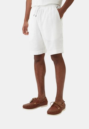 Uomo con pantaloncini bianchi con coulisse e scarpe da barca in pelle marrone, in piedi con una mano nella tasca davanti a uno sfondo semplice.