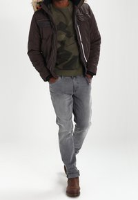 Mand iført brun vinterjakke med pelskrave, grøn camo sweatshirt, grå jeans og brune læderstøvler, gående med hænderne i lommerne.