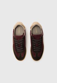 Zapatillas de ante en burdeos y marrón oscuro con acentos en beige, cordones planos marrones y un logo en el interior, con un cuello texturizado en beige.