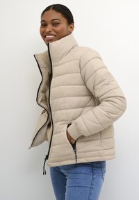 Beige gewatteerde jas met hoge kraag, zwarte ritssluiting, twee zijzakken en elastische manchetten. Gladde textuur met horizontaal quiltpatroon.