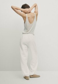 Lichtgrijze mouwloze top met een diepe V-rug, gecombineerd met loose-fitting witte broeken en zachte beige instappers. Minimalistisch ontwerp.