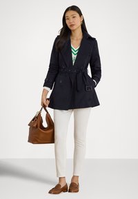 Tmavomodrý dvojradový trenčkot so zúženým pásom, doplnený o biele priliehavé nohavice a hnedé mokasíny. Hnedá kabelka dotvára outfit.