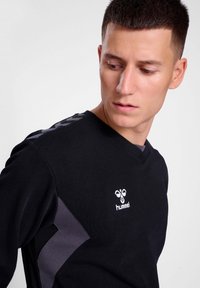 Maglietta nera a maniche lunghe con scollo a V, caratterizzata da accenti grigi sulle spalle e un logo bianco sul petto. Materiale in cotone, texture liscia.