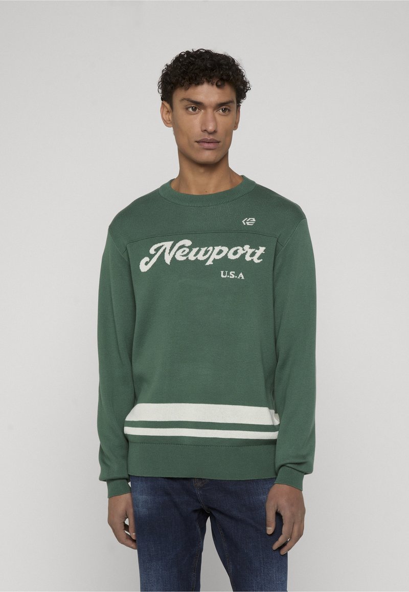 Homme portant un pull en maille vert avec le texte "Newport U.S.A" et des rayures blanches près de l'ourlet, assorti à un jean bleu, debout devant un fond uni.