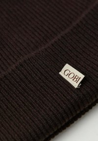 Brun ribbad stickad tyg med en etikett som står "GOBI" i bruna bokstäver på en gräddfärgad bakgrund. Slät textur, synlig vikning vid hörnet.