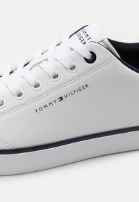 Baskets synthétiques blanches avec bout rond, design plat à lacets, accents bleu marine et logo "TOMMY HILFIGER" imprimé sur le côté.