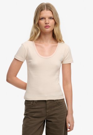 Kvinde med langt blondt hår iført en kortærmet, lysebeige ribbet top og olivengrønne bukser mod en ensfarvet hvid baggrund.