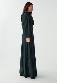 Long dark green dress features ruffled detailing at the neckline, long sleeves, and a tiered skirt. Made of flowing fabric with a smooth texture.