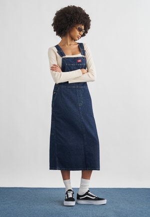 Femme aux cheveux bouclés portant des lunettes de soleil, un haut à manches longues crème, une robe salopette en denim foncé, des chaussettes blanches et des baskets noires, debout les bras croisés.