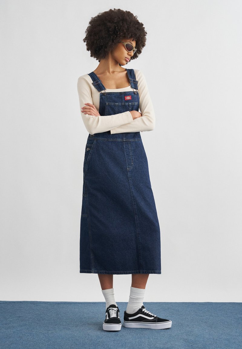 Γυναίκα με σγουρά μαλλιά που φορά γυαλιά ηλίου, κρεμ μακρυμάνικη μπλούζα, σκούρο τζην φόρεμα με τιράντες, λευκές κάλτσες και μαύρα αθλητικά παπούτσια, στέκεται με σταυρωμένα τα χέρια.