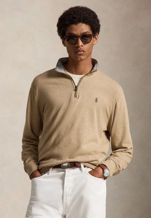 Jeune homme portant des lunettes de soleil foncées, un pull beige à demi-fermeture éclair, un pantalon blanc, une ceinture marron et une montre-bracelet, les mains dans les poches.