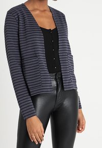 Randig marinblå cardigan med öppen front, två fickor och avslappnad passform, kombinerad med en svart ribbad topp och skinande svarta leggings.