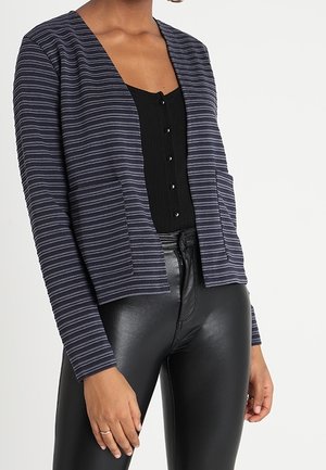 Cárdigan de rayas azul marino con front abierto, dos bolsillos y un corte relajado, combinado con una blusa negra de canalé y leggings negros brillantes.