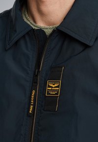 Marineblå vindjakke med lynlås foran, der har et tekstureret mærke med gule accenter, der står "PME LEGEND" og "AMERICAN CLASSIC."