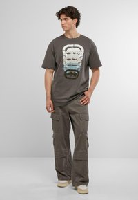 Grå bomulds T-shirt med et flerfarvet grafisk design, der viser bjørnsilhuetter, parret med grå cargobukser og beige sneakers.