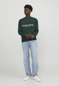 Sudadera verde con un logo en blanco y naranja, combinada con jeans de un azul claro y zapatillas blancas. La tela parece suave y casual.