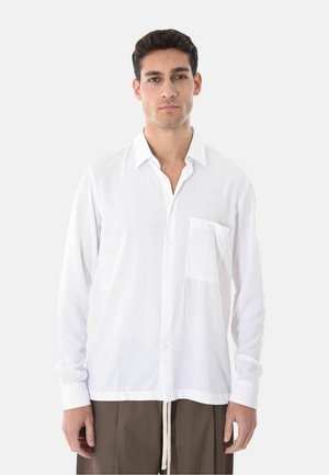 Uomo con capelli corti e scuri che indossa una camicia bianca a maniche lunghe abbottonata e pantaloni marroni, in piedi su uno sfondo chiaro e uniforme.