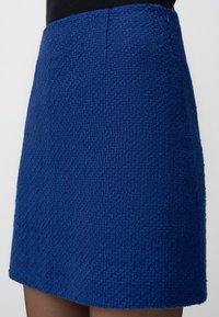 Jupe texturée bleue avec un ourlet court, présentant une finition lisse et une fermeture éclair sur le côté. Le tissu a une apparence tissée.