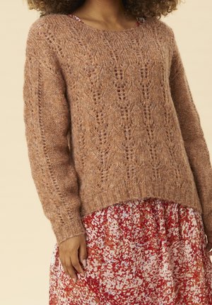 Pull à torsades marron porté sur une robe fleurie rouge, blanche et marron sur une personne aux cheveux bouclés sur fond beige.