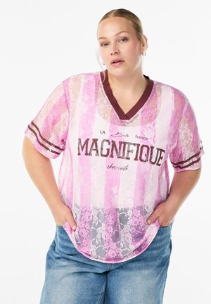 Pink and white lace short-sleeve top with a V-neck, featuring "MAGNIFIQUE" text and horizontal stripes on sleeves, paired with blue jeans.