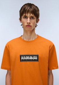 Orange bomulds t-shirt med korte ærmer, prydet med et sort rektangulært logo med hvid tekst i midten. Glat tekstur, rund halsudskæring.