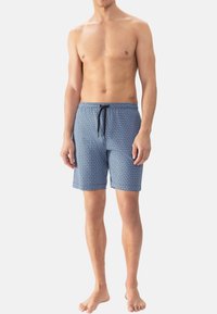 Man staat blootvoets in blauwe gestreepte badshorts met een zwarte drawstring tegen een plain witte achtergrond.