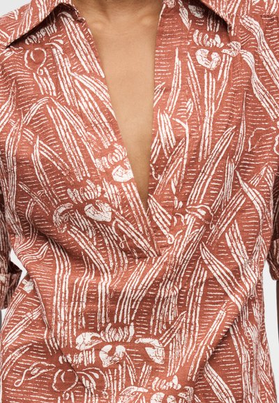 Chemise décolletée en V profond avec une base rouille et un motif botanique blanc comprenant des feuilles et des fleurs, montrant partiellement la poitrine et le col.